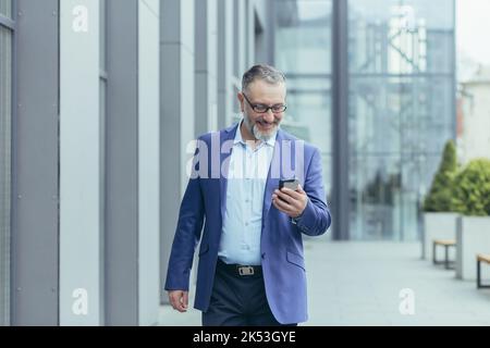 Senior gray-haired handsome man in a suit and glasses. Walks down the street, holds the phone in his hands, makes a video call, talks. Stock Photo