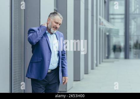 An senior man, a banker, an office worker in suit, went outside the office for a lunch break, his neck hurts, he massages his neck with one hand, he feels pain. Stock Photo