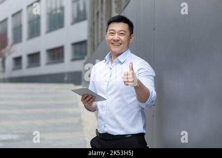 Portrait of a young handsome asian man in a white shirt standing on the street near a modern building. Holds a tablet in his hand, types, works. Shows super. He looks at the camera, smiles. Stock Photo