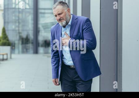 Pre-infarction condition. A senior man in a suit stands on the street near an office center holding his heart. He feels a sharp pain in his chest. He needs urgent medical help. Stock Photo