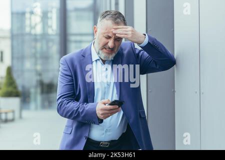 A senior man, a businessman, is standing on the street near an office center, holding his head in his hand, having a severe headache. Calls an ambulance by phone. Stock Photo