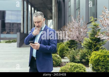 Pensive and worried senior man in a business suit stands on the street near a modern building, looks at a mobile phone, reads, types, holds his beard thoughtfully. Stock Photo