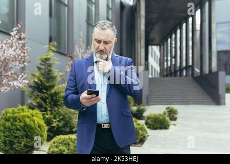 Pensive and worried senior man in a business suit stands on the street near a modern building, looks at a mobile phone, reads, types, holds his beard thoughtfully. Stock Photo