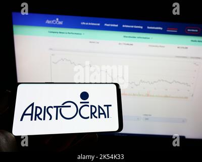 Person holding mobile phone with logo of gambling company Aristocrat Leisure Limited on screen in front of web page. Focus on phone display. Stock Photo