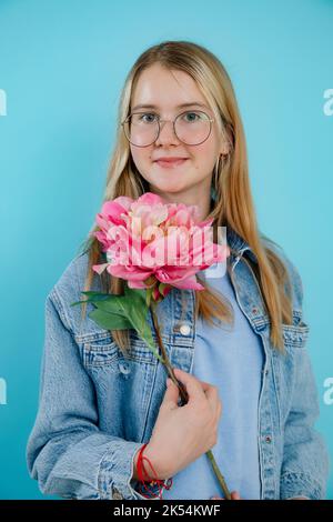 Cute teenage girl holding big fragrant pink fresh flower with long stem on empty blue background. Happy young lady in glasses with smile enjoy beauty Stock Photo