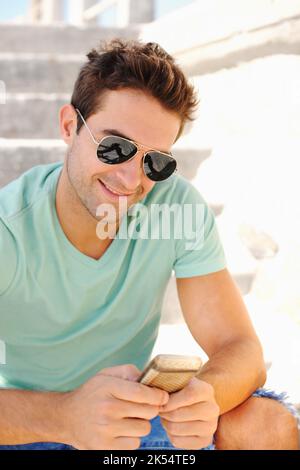 Taking time to text. A handsome young man wearing sunglasses and smiling while seated on a flight of stairs and texting on his cellphone. Stock Photo