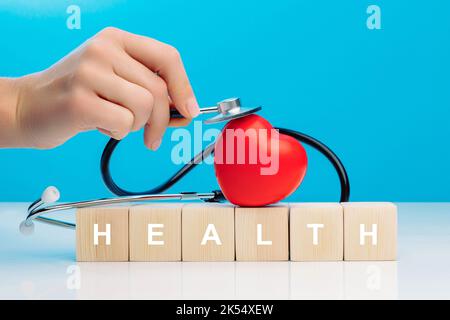 A woman's hand holds a stethoscope and touches a red heart that lies on top of wooden cubes with the inscription HEALTH Stock Photo