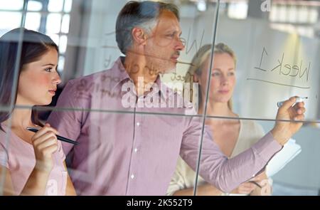 Setting up a schedule for their latest project. A mature businessman writing down plans on a glass pane while his colleagues look on. Stock Photo