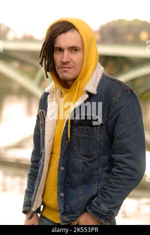 Portrait of young positive man with dreadlocks in hood, posing in city park on background of blurred bridge in cloudy weather. Stock Photo