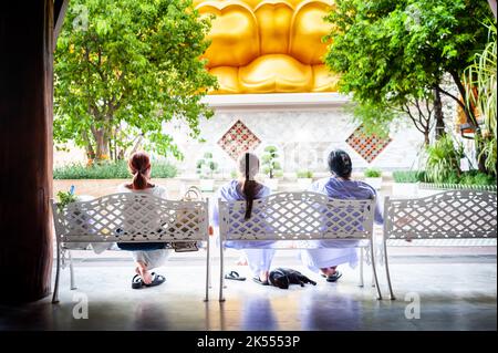 The giant golden Buddha looms large above three Thai ladies sat peacefully at a bench at Wat Paknam. A black cat sleeps at their feet. Stock Photo