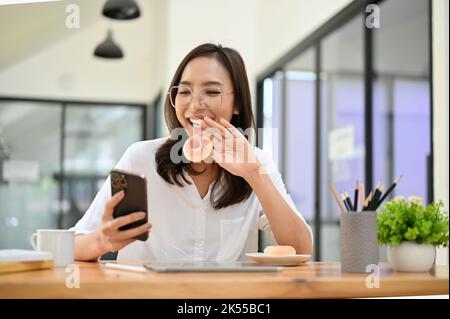 Beautiful and happy young Asian female office worker wearing eyeglasses, sits at her office desk, enjoys eating doughnuts while chatting with her frie Stock Photo