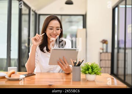 Attractive and happy young Asian businesswoman or female office worker wearing eyeglasses using digital tablet while remote working in the cafe. Stock Photo
