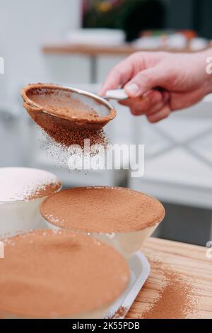 The Process of preparing tiramisu dessert. The dish is sprinkled with cocoa through a sieve. Close-up, selective focus. Stock Photo