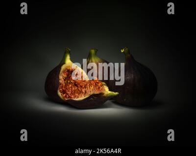 Three black Turkish figs, one split to show seeds. Dark still life, chiaroscuro. Stock Photo