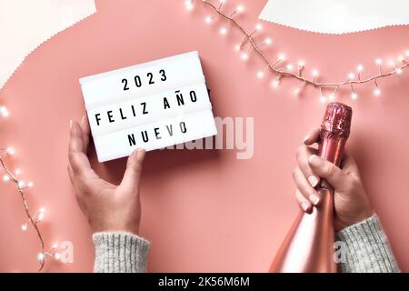 Text Happy New Year 2023 in Spanish Language on lightbox. Light garland, woman hands with lightbox and metallic pink champagne bottle. New Year Stock Photo
