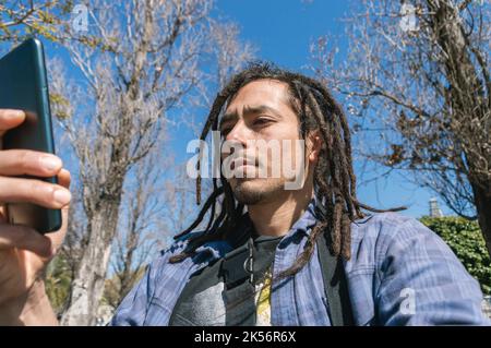 young latin venezuelan man with dreadlocks, and a blue plaid shirt is outdoors, he is holding his phone with one hand worried reading a text message Stock Photo