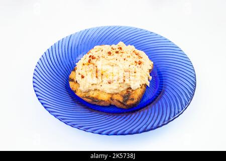Humus paste with paprika powder on a toasted slice of bread, on a blue glass plate, isolated on white background. Side view close up. Stock Photo