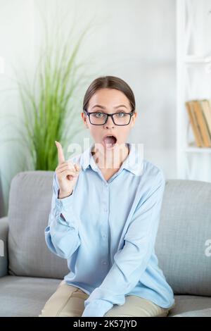 Smart idea. Aha insight. Remember recommendation. Inspired confident woman in eyeglasses with open mouth pointing up on couch at light home interior w Stock Photo