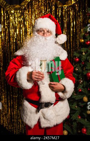 Santa claus holding glass of champagne and gift near christmas tree and tinsel,stock image Stock Photo