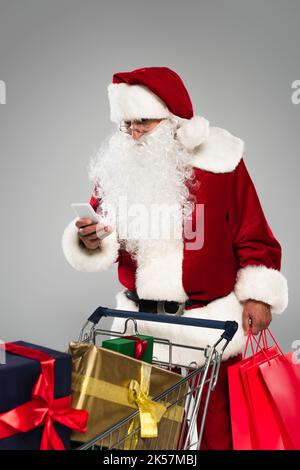 Santa claus using mobile phone and holding shopping bags near gifts in cart isolated on grey,stock image Stock Photo