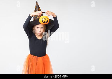 A cheerful little girl in a carnival witch costume and hat, holding a basket for treats, scares on a white background. Children's Halloween. Trick or Stock Photo