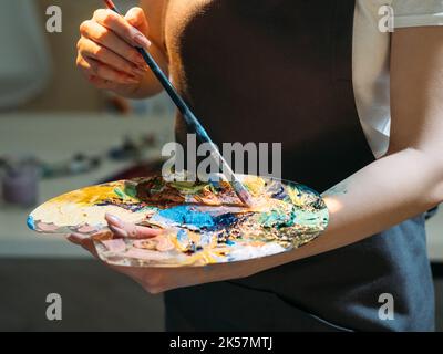 Mixing colors. Female artist. Creative process. Art tools. Unrecognizable woman blending colorful paints on palette with brush in light studio interio Stock Photo