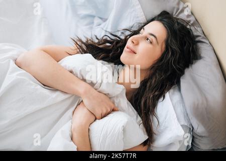 Romantic morning. Weekend rest. Pleasure wellbeing. Peaceful thoughtful daydreaming brunette woman smiling enjoying lying in cozy bed with white soft Stock Photo