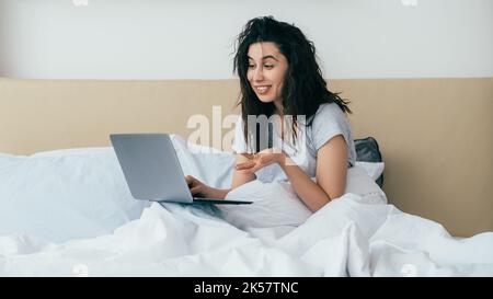 Morning online. Video chat. Digital meeting. Web communication. Surprised curious happy woman speaking discussing life using laptop smiling in bed in Stock Photo