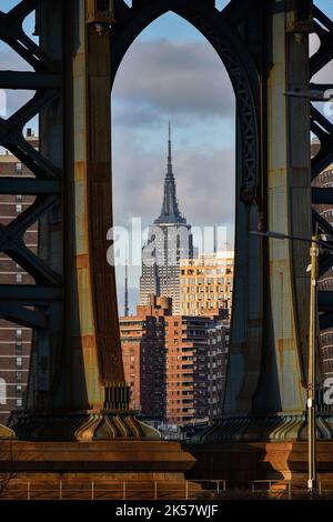 View of the empire state building surrounded by buildings through a pylon of the Manhattan bridge at sunset. Stock Photo