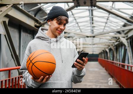 Young man basketball player with headphones holding ball using smartphone after training. Urban background. Stock Photo