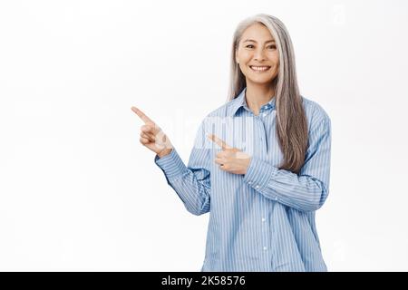 Smiling asian woman looking pleased, showing advertisement, pointing left, standing against white background Stock Photo