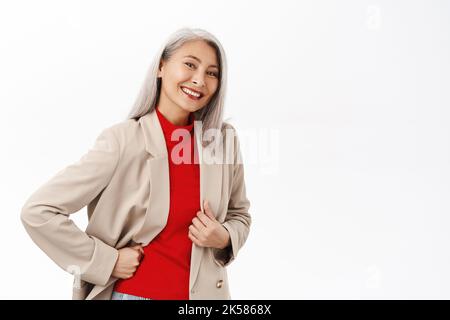 Portrait of healthy and happy senior asian woman in stylish blazer, smiling and looking confident at camera, white smile, standing over white Stock Photo