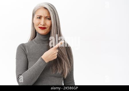 Portrait of confused asian senior woman frowning, pointing right at smth strange, showing promo offer, standing over white background Stock Photo