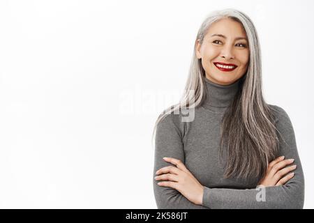 Portrait of beautiful, healthy smiling middle aged asian woman, cross arms on chest, looking confident and happy, standing over white background Stock Photo