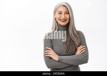 Portrait of beautiful, healthy smiling middle aged asian woman, cross arms on chest, looking confident and happy, standing over white background Stock Photo