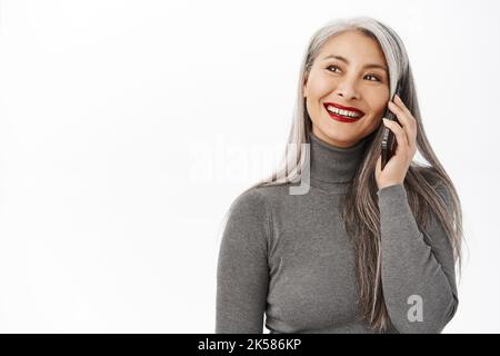 Portrait of beautiful and happy asian senior woman, middle aged lady talking on mobile phone, calling someone and smiling, standing over white Stock Photo