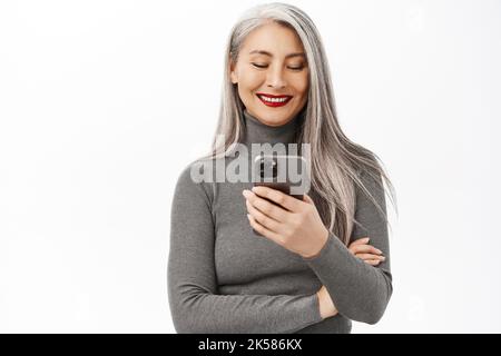 Portrait of beautiful middle aged asian woman holding smartphone and smiling at camera, using mobile app, standing over white background Stock Photo