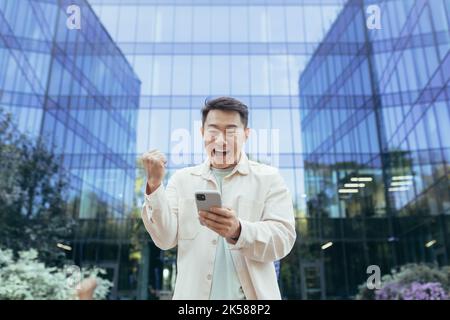 Happy asian man celebrating victory and success holding phone smiling and happy, programmer outside modern office building software company, man in casual clothes using smartphone. Stock Photo