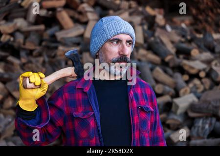 Brunette handsome man with beard holding axe and angry face.  Wearing lumberjack shirt. Wooden blurry background. Stock Photo