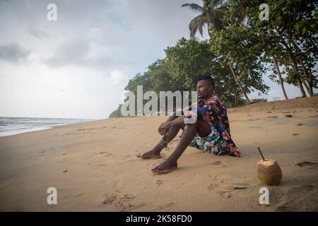 Young man sitting pensively on the beach sand holding his sunglasses enjoying his life, copy space on the left. Stock Photo