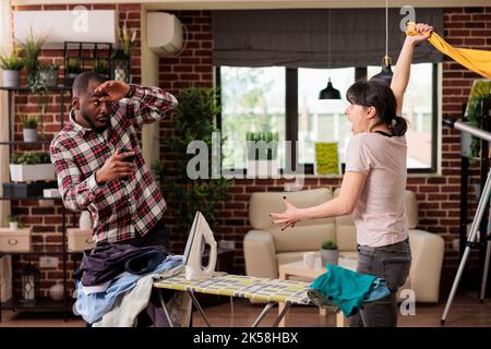 Nervous upset woman trying to hit husband while having a big fight at home. Multiracial couple arguing because tired wife takes care of household chores without help, marital problems. Stock Photo
