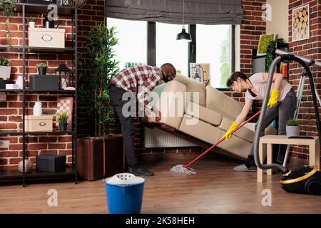 Married couple doing spring cleaning at home, wife wetting the dust under the sofa while husband lifts it up. Multiracial family spending weekend doing household chores. Stock Photo