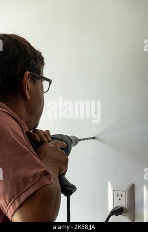 latin man in his 50s, drilling a wall with a drill, white wall Stock Photo