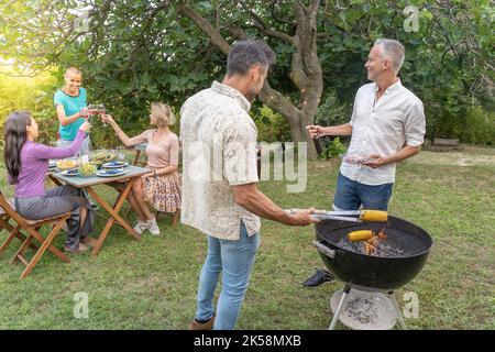 Friends cooking with the barbecue having a conversation smiling happy and laughing together talking in the patio. Stock Photo