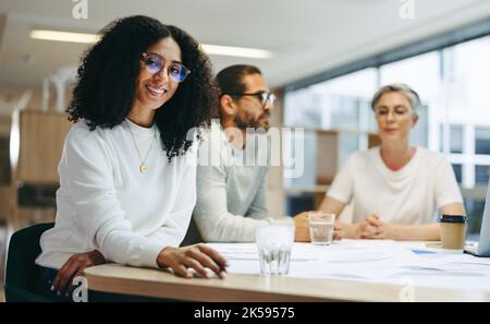 Cheerful young businesswoman smiling at the camera while sitting in a meeting with her team. Group of innovative design professionals sharing creative Stock Photo