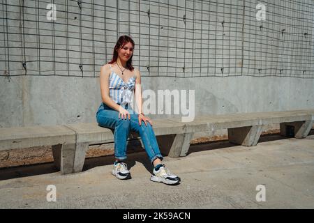 Beautiful latin woman with red hair sitting on a cement bench wearing sunglasses and wearing a jean and a sleeveless top on a sunny day. Copy space. L Stock Photo