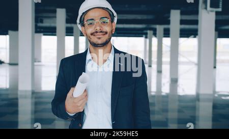 Portrait of cheerful Arab man architect holding blueprint and wearing helmet standing indoors empty building smiling and looking at camera Stock Photo