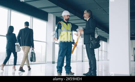 Construction supervisor mature man in suit is talking on mobile phone then discussing project with foreman guy in uniform vest and helmet. Stock Photo