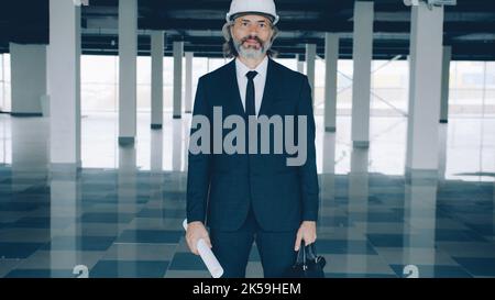 Portrait of experienced chief engineer holding blueprint and wearing helmet standing in modern plant building alone ready for real estate deal Stock Photo