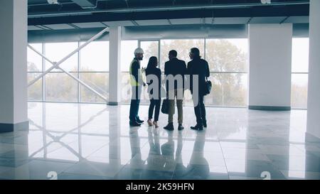 Group of engineers male and female are discussing new building interior checking blueprint and talking standing against panoramic window background Stock Photo